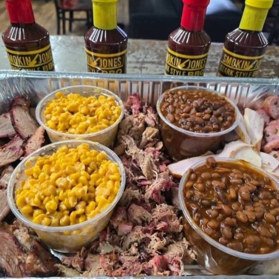 Smokin' Jones BBQ platter with pulled pork, sliced meats, baked beans, and corn, with sauce bottles.