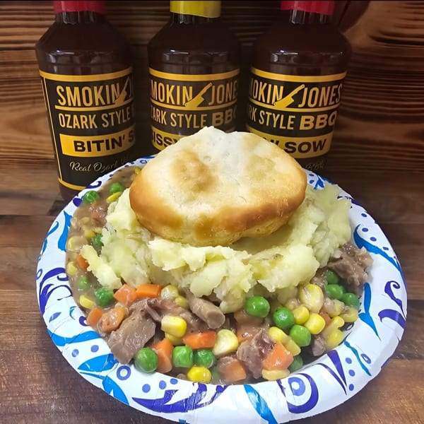 Smokin' Jones BBQ stew with mashed potatoes and a biscuit, with three BBQ sauce bottles in the background.