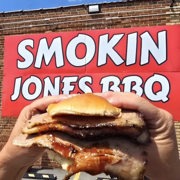 Hands holding a saucy brisket sandwich in front of the Smokin' Jones BBQ sign.