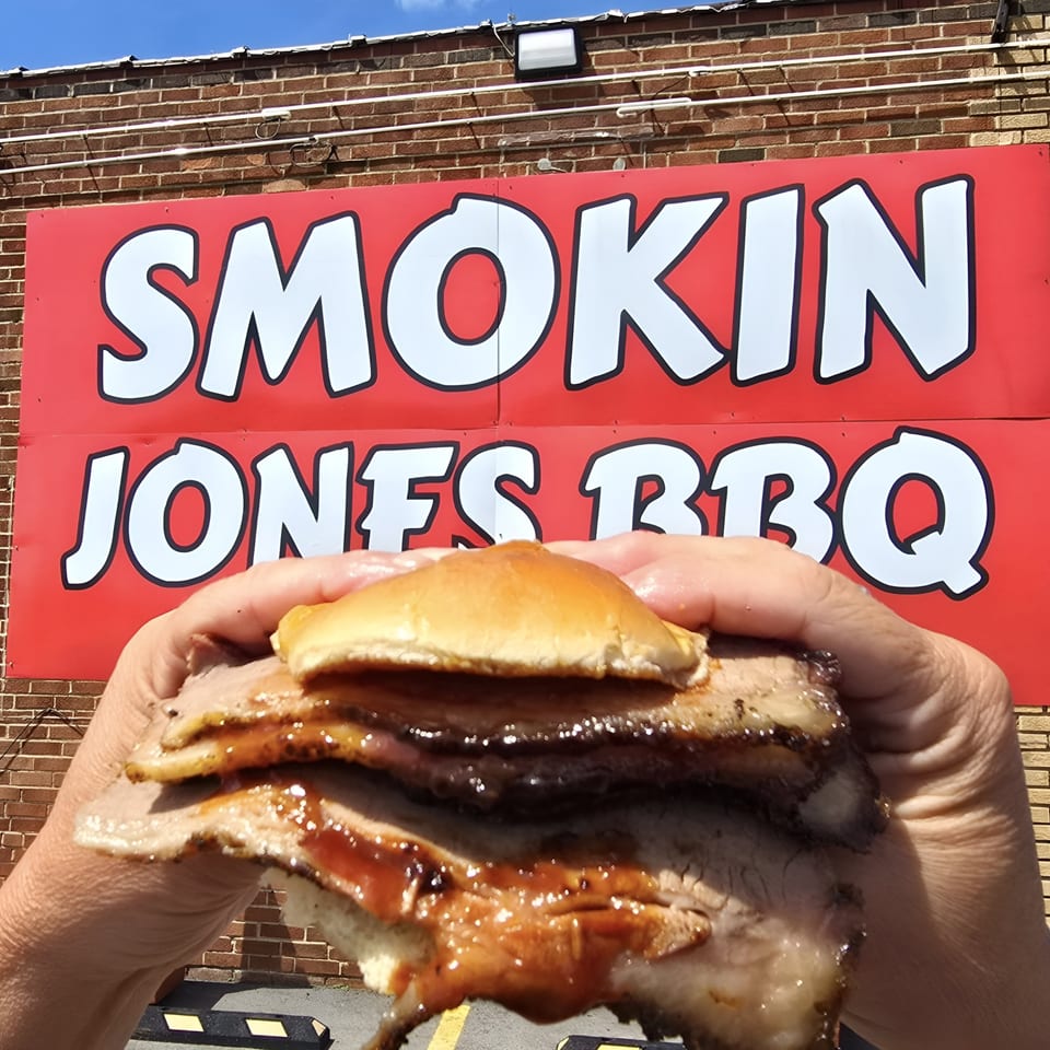 Hands holding a saucy brisket sandwich in front of the Smokin' Jones BBQ sign.
